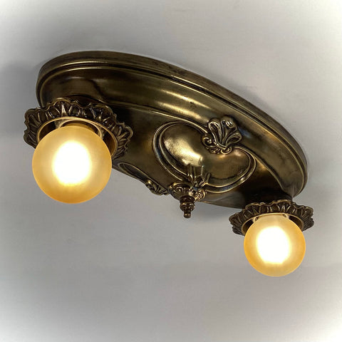 Antique brass two-light flush mount ceiling fixture with illuminated frosted amber bulbs, bottom view.