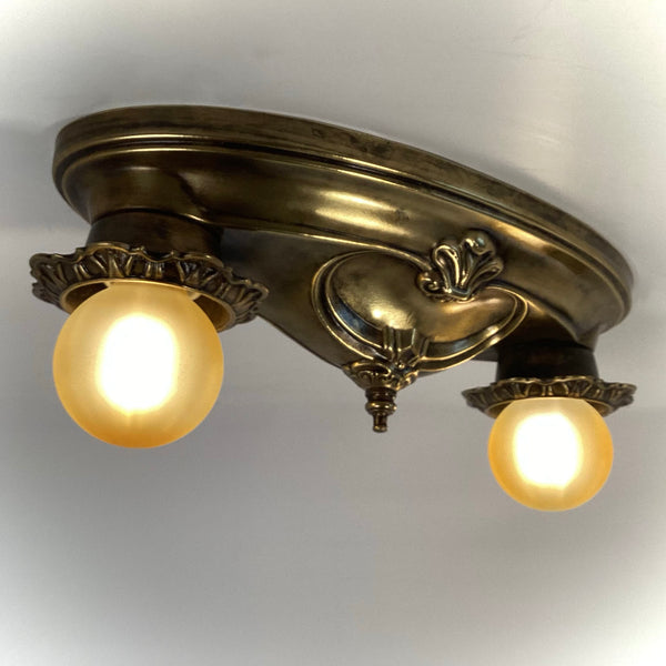 Angled view of an antique brass two-light ceiling fixture with warm illuminated globes.