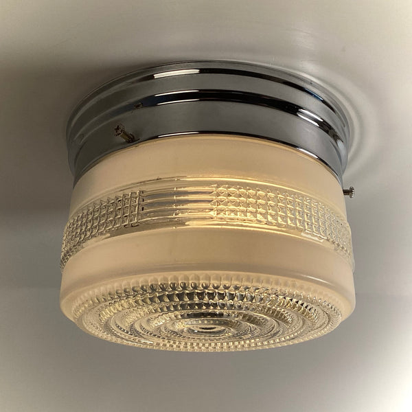 A close-up, angled view of an illuminated vintage Art Deco flush mount ceiling light with a textured glass shade and polished chrome fixture.