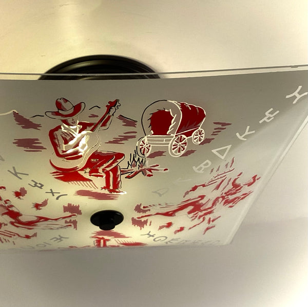 Detailed close-up of a vintage glass light shade depicting a cowboy playing guitar next to a campfire and covered wagon in red print.