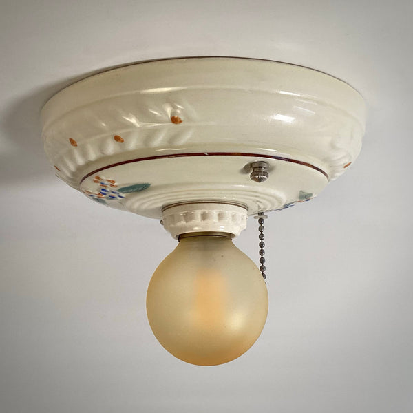An antique porcelain flush mount ceiling light with hand-painted floral details, showing a dimmed, softly glowing round bulb.