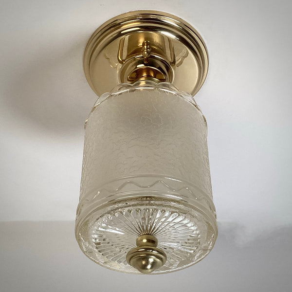 Polished brass semi-flush mount light fixture with textured glass cylinder shade, shown unlit against a white ceiling.