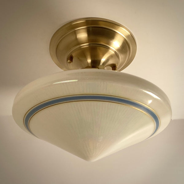 Low-angle view of a vintage Art Deco schoolhouse flush mount light fixture showcasing the sleek satin brass hardware and blue-striped glass shade.