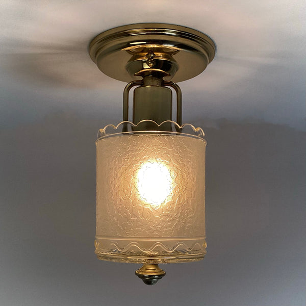 Eye-level lit view of a vintage brass ceiling light featuring a textured clear glass shade and decorative brass finial.