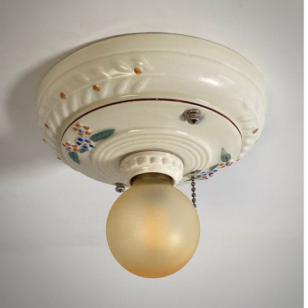 An angled view of a Depression-era vintage ceramic flush mount light fixture featuring blue and orange painted flowers.