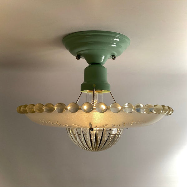 Profile view of an illuminated vintage glass ceiling light with a green flush mount base and clear beaded rim.