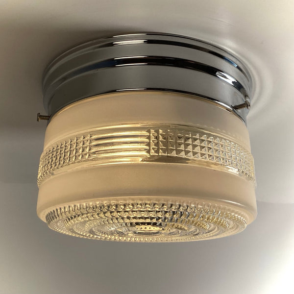 An angled side view of a turned-on retro chrome flush mount ceiling light, emitting a warm golden glow through its frosted and textured glass.