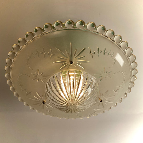 Looking up at an illuminated vintage frosted glass ceiling fixture with etched starburst patterns and a textured center dome.