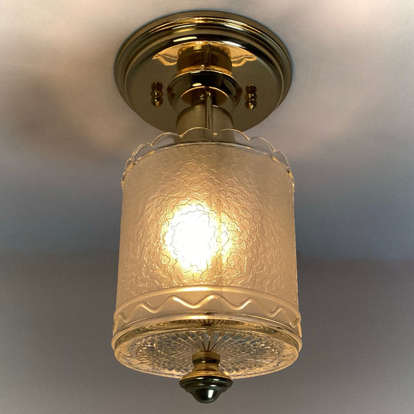 Illuminated view of a vintage brass ceiling light highlighting the canopy mounting hardware and textured glass shade.