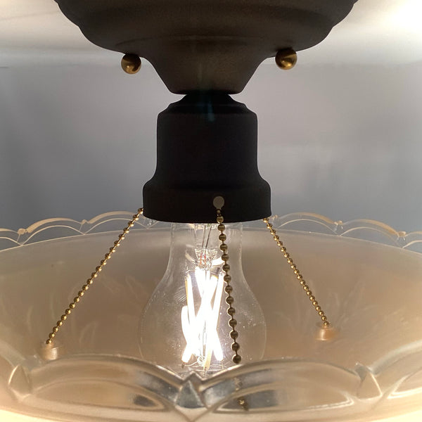 Close-up of a dark metal ceiling socket with exposed LED filament bulb, brass mounting screws, and ball chain shade supports.